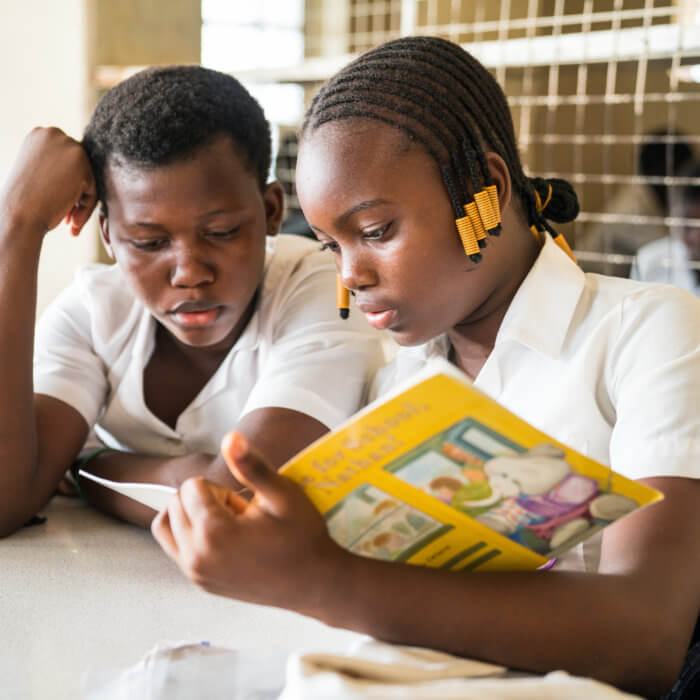 Sarah James (left), 13 years old, and Bintu Rabie Conteh (right), 12, are enrolled in Rising Academy Network's Tengbeh Town school as JSS1 students. Bintu is helping Sarah, a 